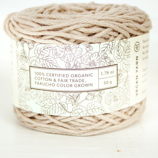 A skein of beige cotton yarn with a label indicating it is 100% certified organic, fair trade, and color grown.