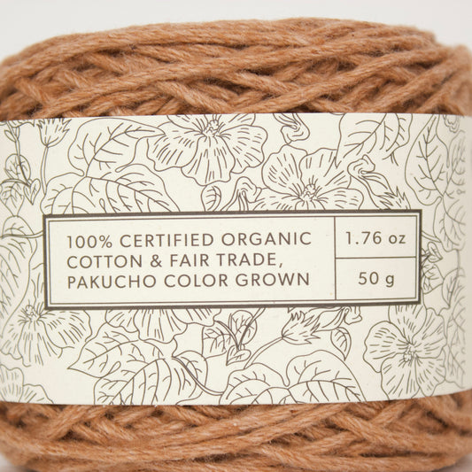 A ball of Pakucho color grown cotton yarn with a white paper label featuring black floral line art. The label text reads '100% CERTIFIED ORGANIC COTTON & FAIR TRADE, PAKUCHO COLOR GROWN' with weight details '1.