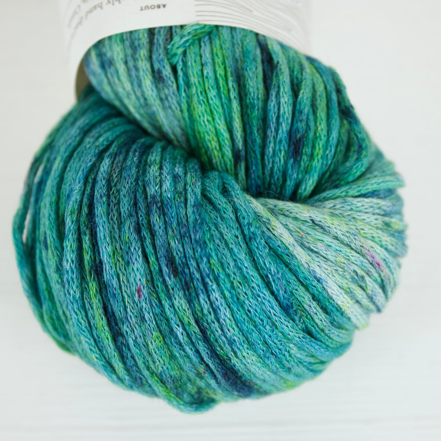 Close up of a skein of Rainy Pond colourway on Taika Aran. It's a speckled yarn with lots of speckles, so the yarn is saturated with grassy greens, indigo, sky blue, aqua, royal blue, and the occasional magenta fleck. 