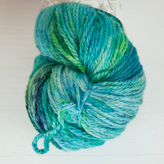 Close up of a skein of Rainy Pond colourway. It's a speckled yarn with lots of speckles, so the yarn is saturated with grassy greens, indigo, sky blue, aqua, royal blue, and the occasional magenta fleck. 