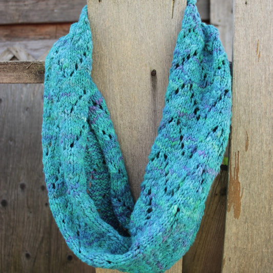 teal cowl with purple flecks, knitted flat with a chevron or zigzag pattern of eyelets that's joined in a circle from end to end. The cowl is hanging on a wooden fencepost, and there's a wooden shed in the background.